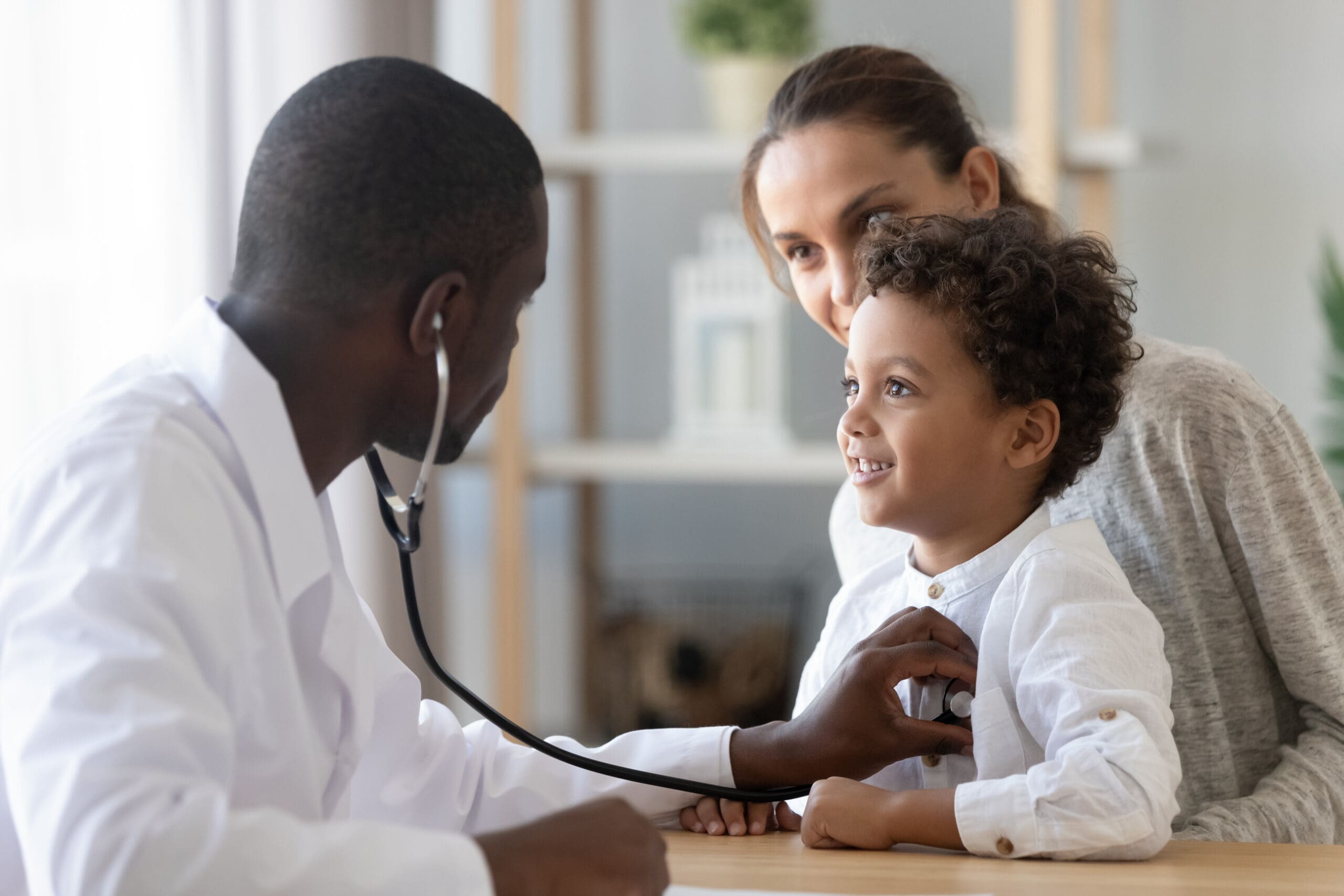 African American male pediatrician with stethoscope listening to lung and heart sound of little boy sitting on mother's lap